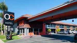 Un hôtel au design moderne, avec une enseigne bien visible arborant le logo et le nom de l’établissement, un parking couvert et un bâtiment à deux étages aux couleurs rouge et grise.