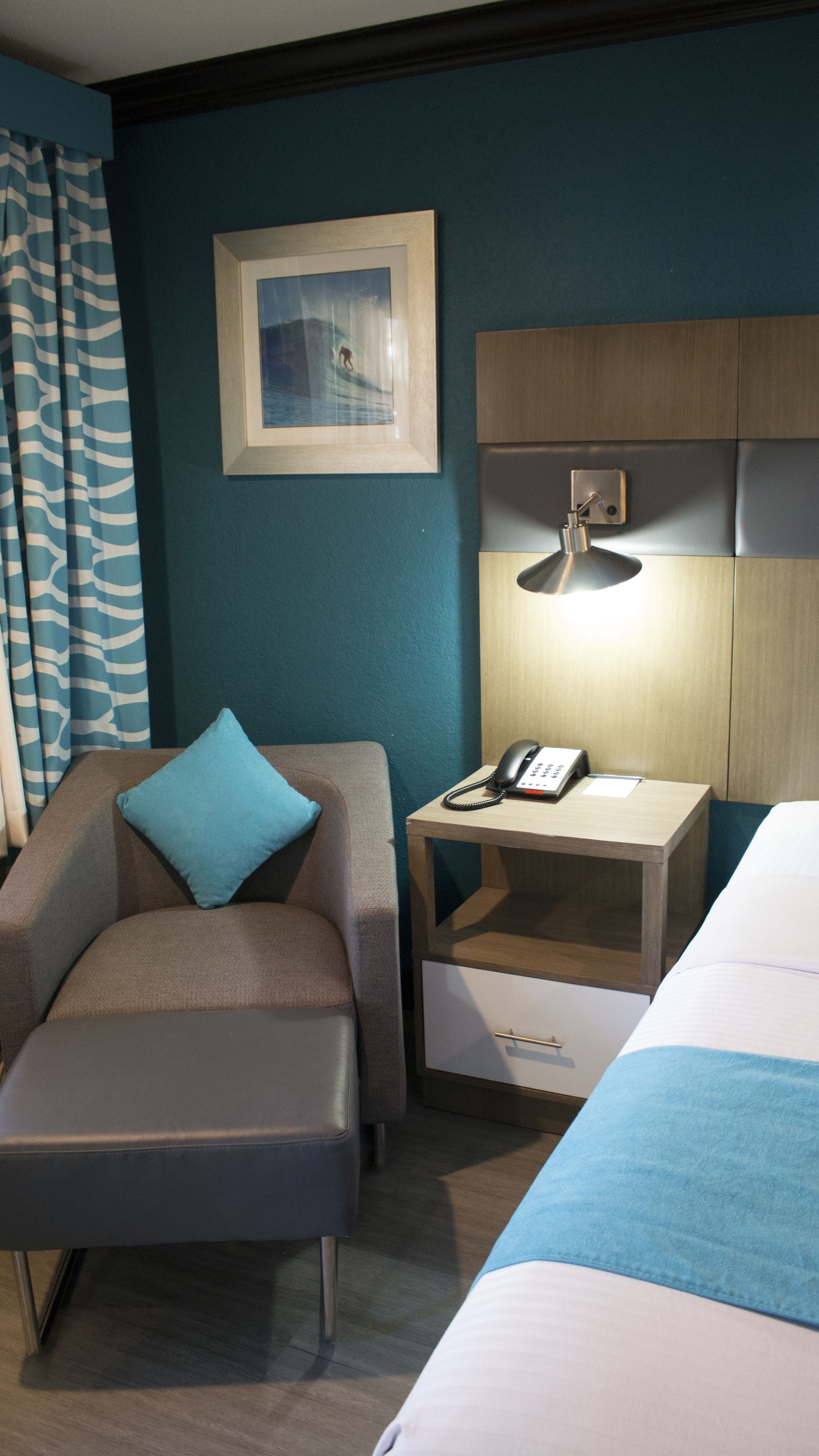 Une chambre d’hôtel avec un lit, un bureau avec un téléphone, une chaise avec un coussin, un tableau encadré au mur et un rideau bleu.