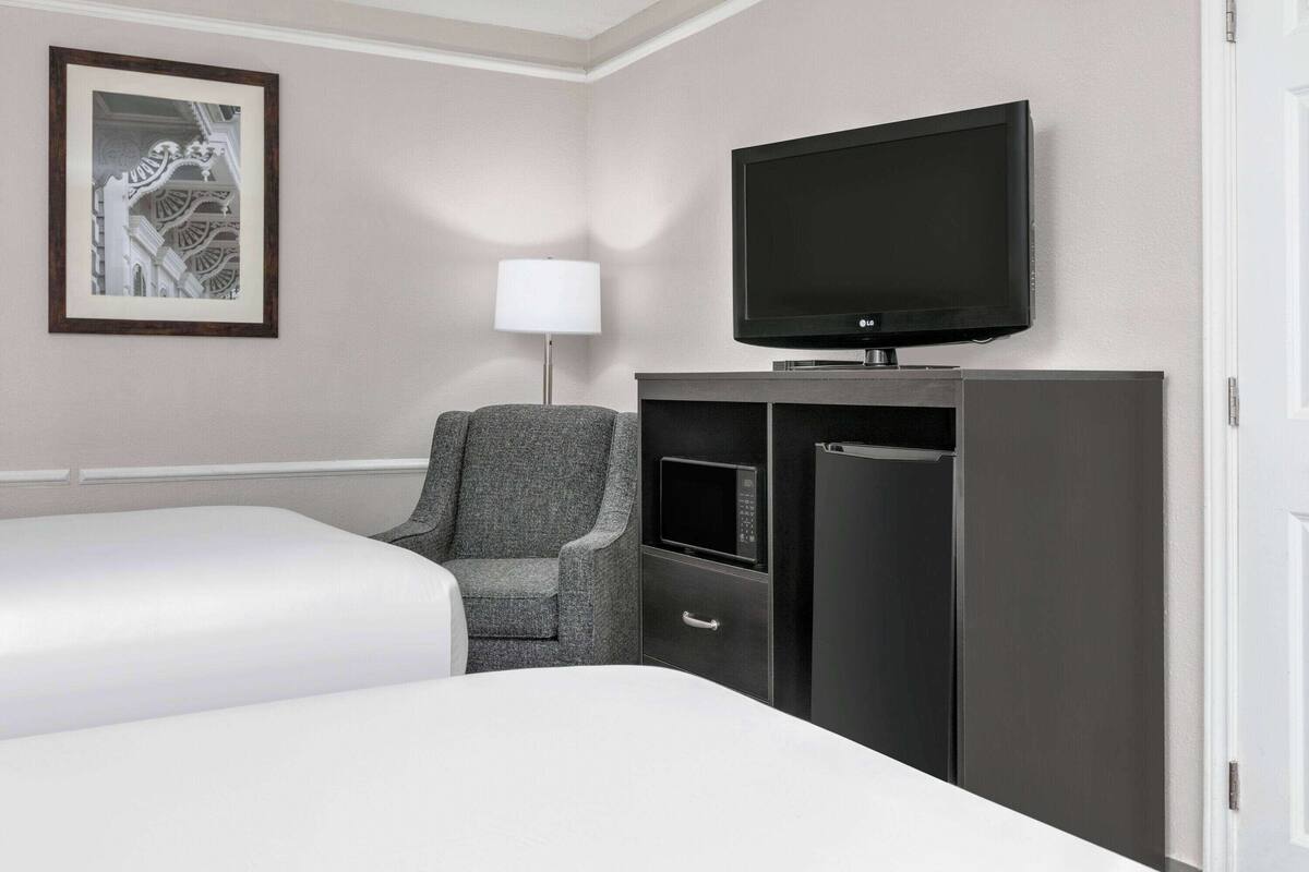 A hotel room with a bed, a gray armchair, a flat-screen TV, a microwave, and a minibar.