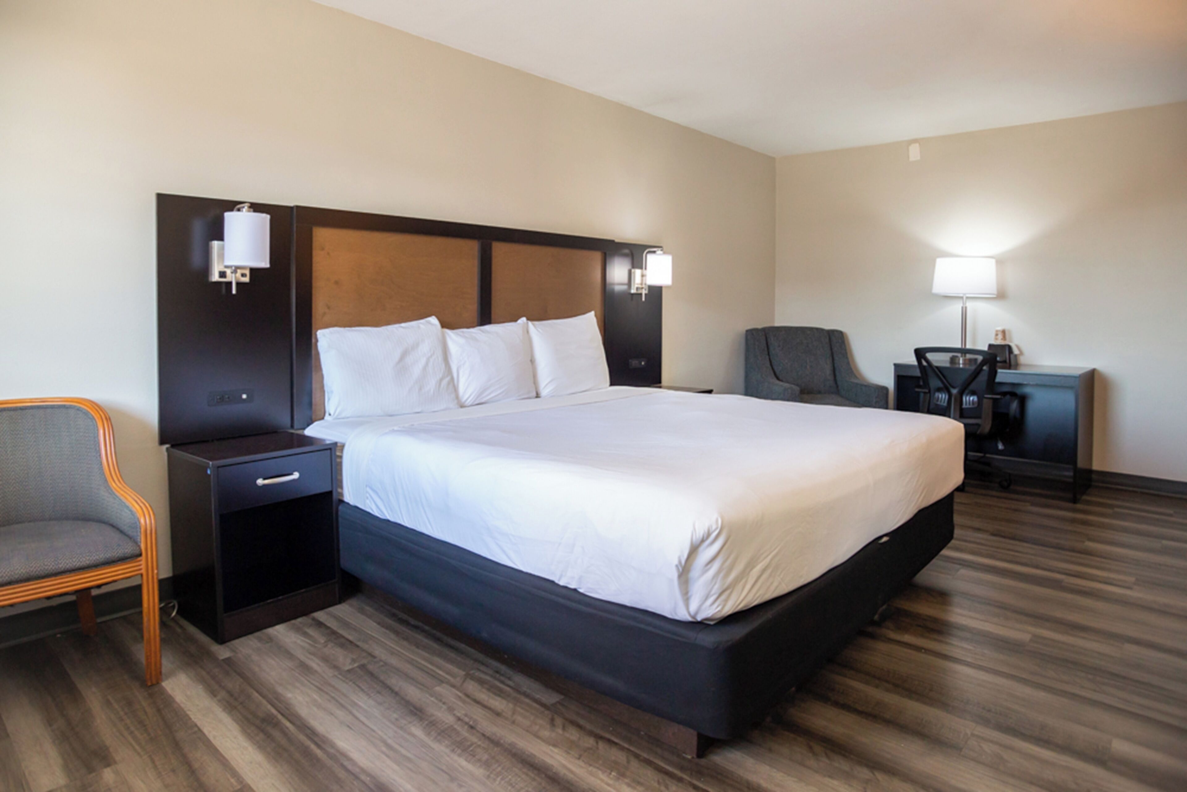 A hotel room with a large bed, a nightstand, a chair, and a desk with a lamp.