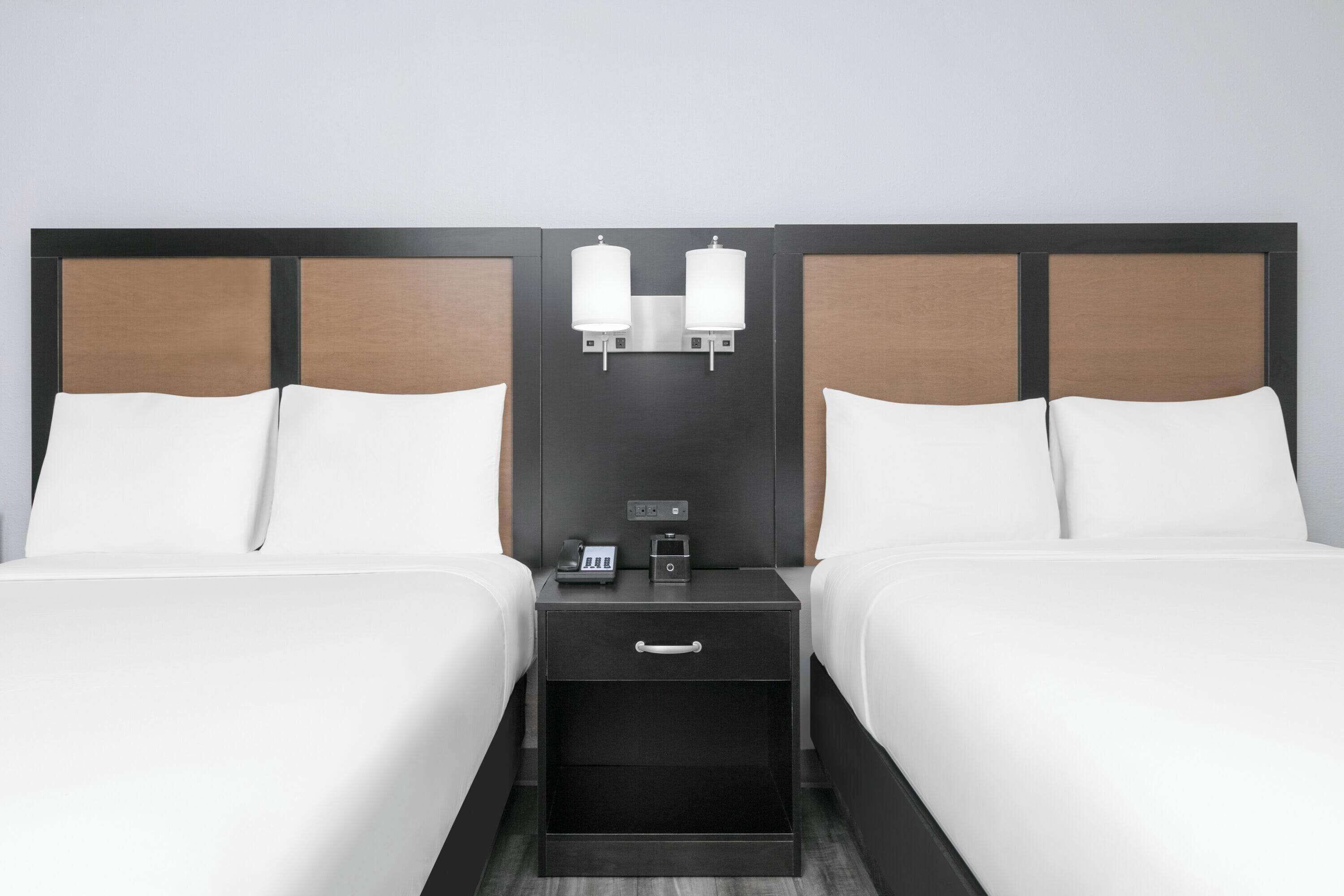 A hotel room with two beds, a nightstand with a phone and a small table, and wall-mounted lamps.