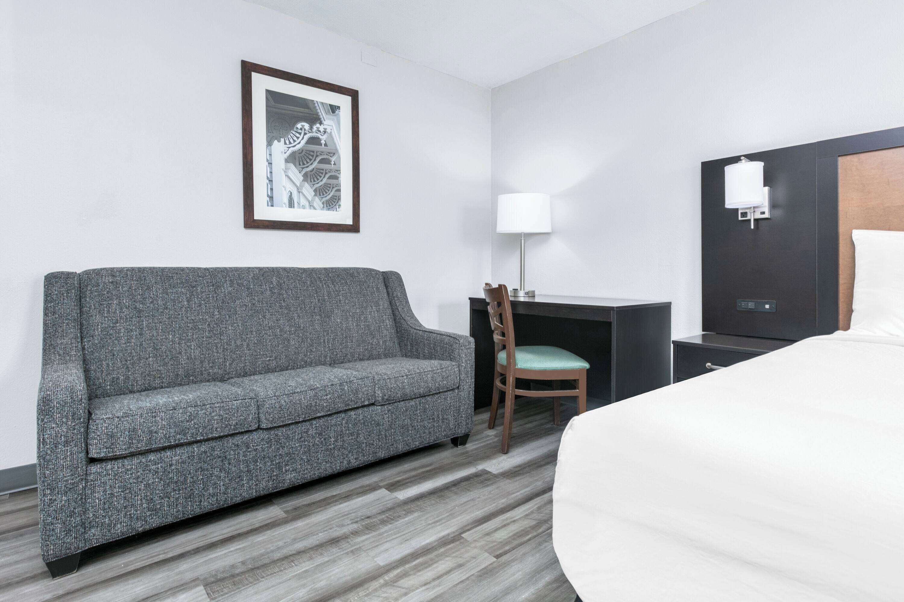 Ein Hotelzimmer mit grauem Sofa, einem Holzstuhl, einem Schreibtisch mit Lampe, einem Bett und einem gerahmten Bild an der Wand.