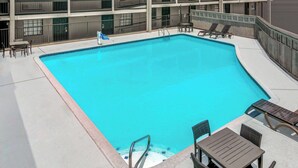 Une piscine rectangulaire à l’intérieur bleu, entourée d’une terrasse blanche. On y trouve des chaises longues et une table avec des chaises sur la terrasse.