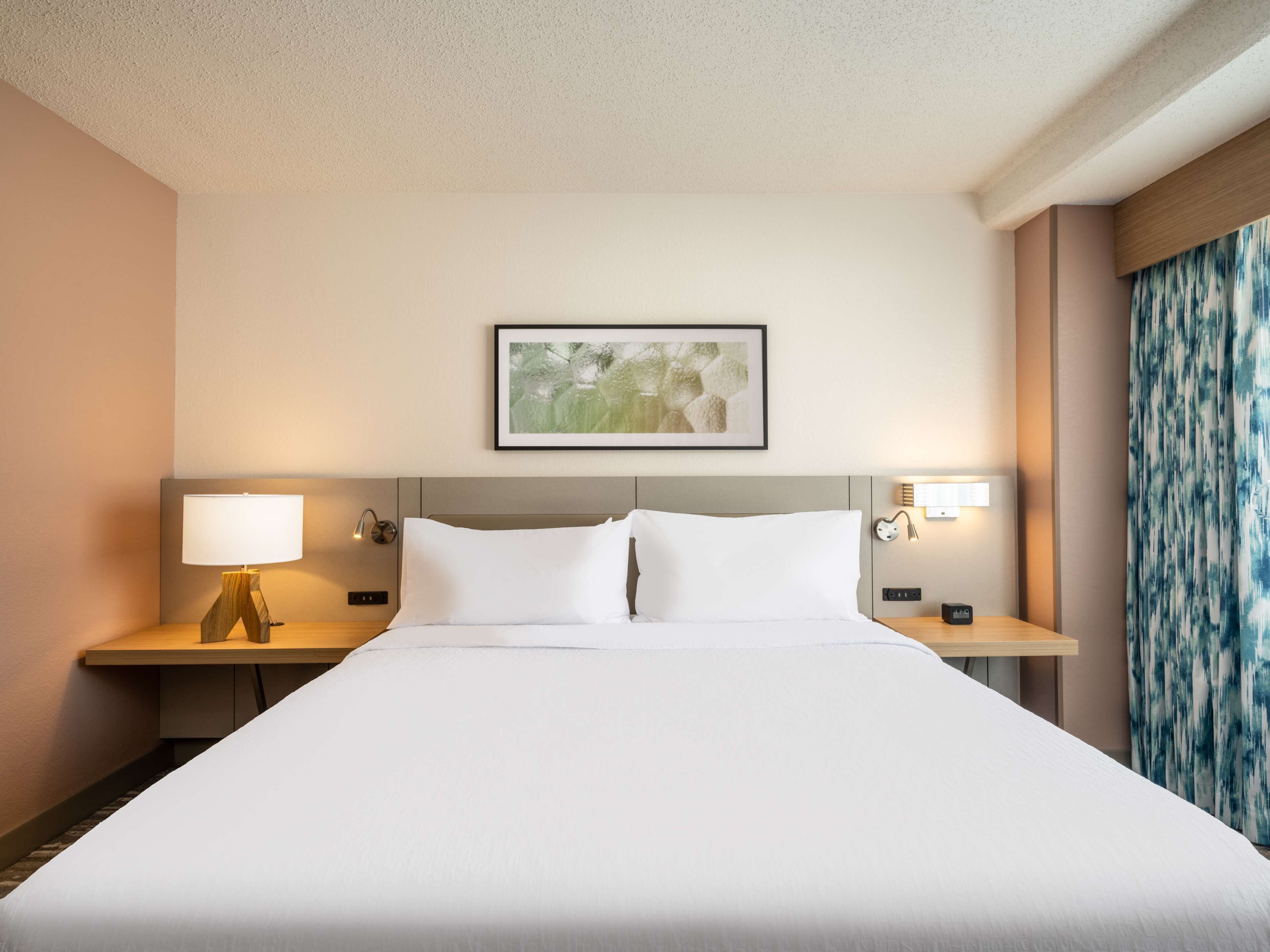 Habitación de hotel con una cama grande, dos mesitas de noche con lámparas, una obra de arte enmarcada sobre la cama y una cortina estampada a la derecha.