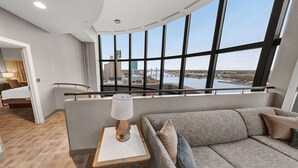 A modern hotel room with a large window offering a city view, a cozy seating area, and a staircase leading to an upper level. - Hilton Garden Inn Toledo Downtown (Toledo)