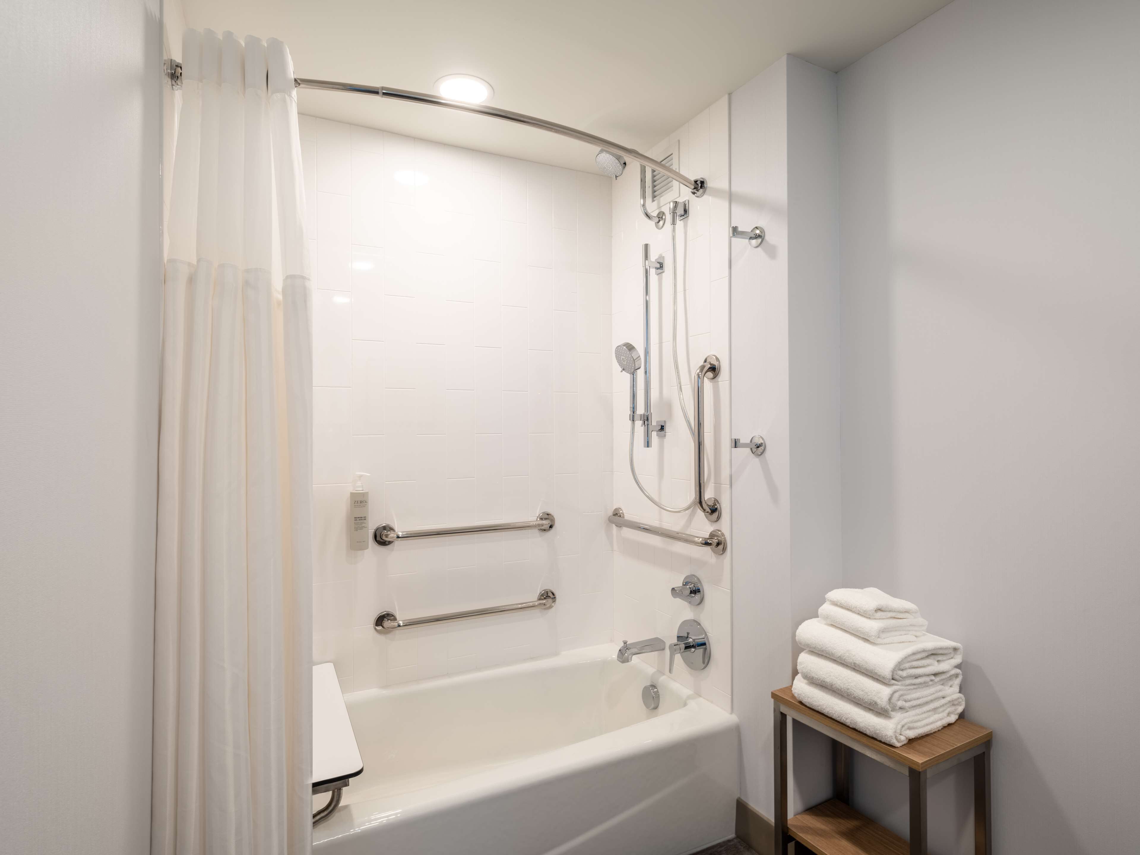 A modern bathroom with a walk-in shower, grab bars, a bathtub, and a stack of towels on a wooden table.