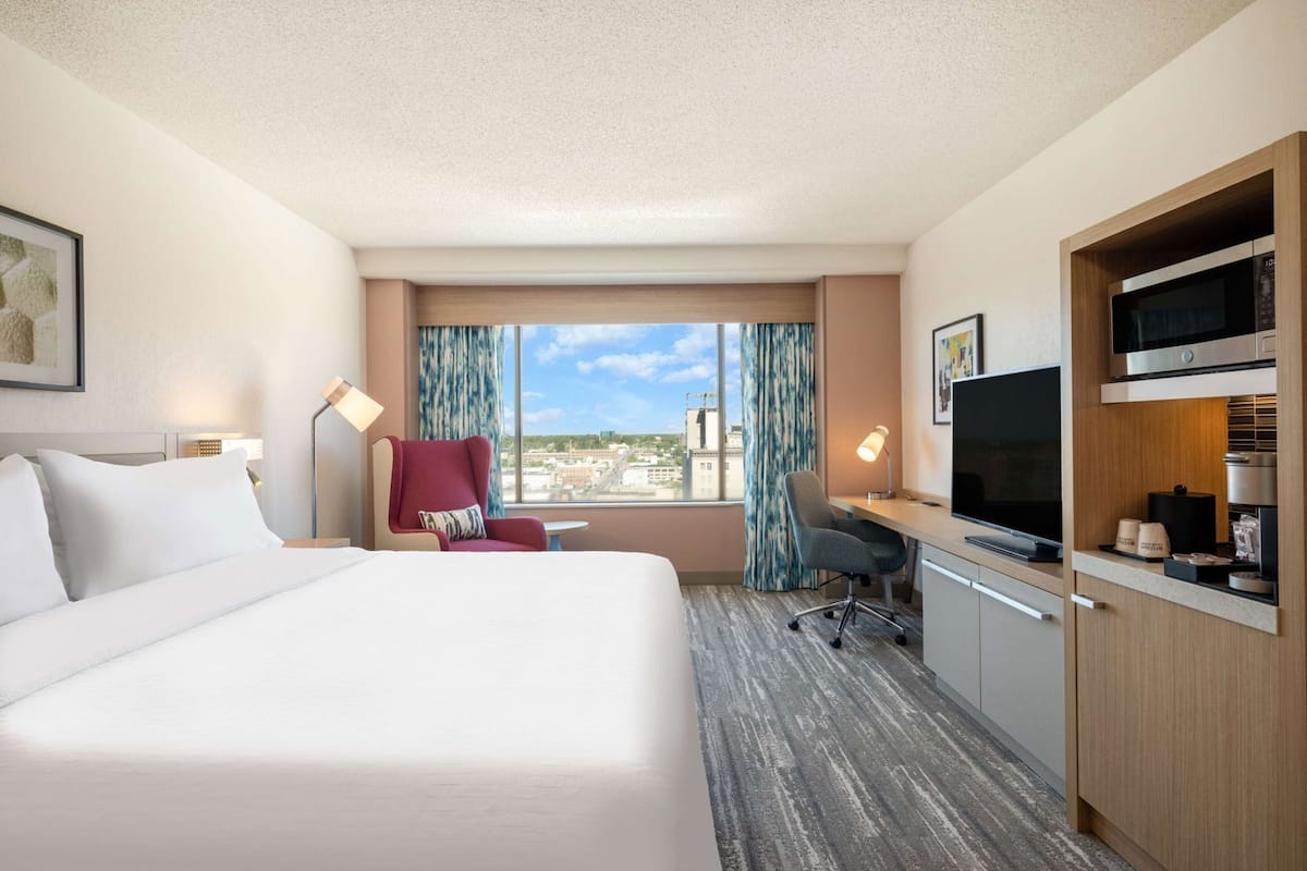 A hotel room with a bed, desk, TV, and a view of the city.