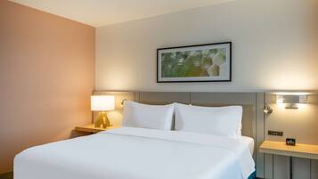 A hotel room with a large bed, a nightstand with a lamp, a wall-mounted artwork, and a bedside table with a clock.