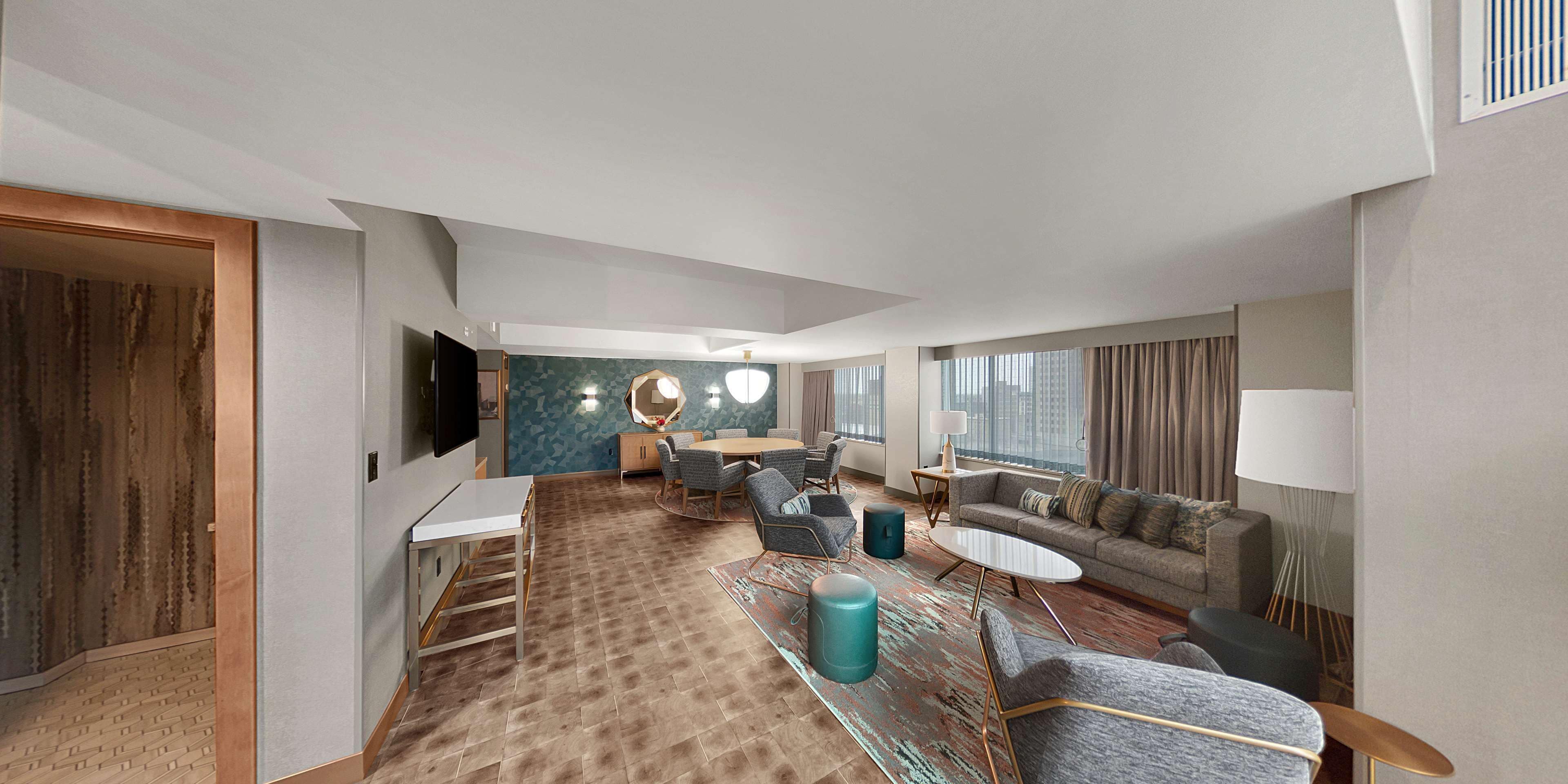 A modern hotel room with a dining area, a sofa, a TV, and a large window.
