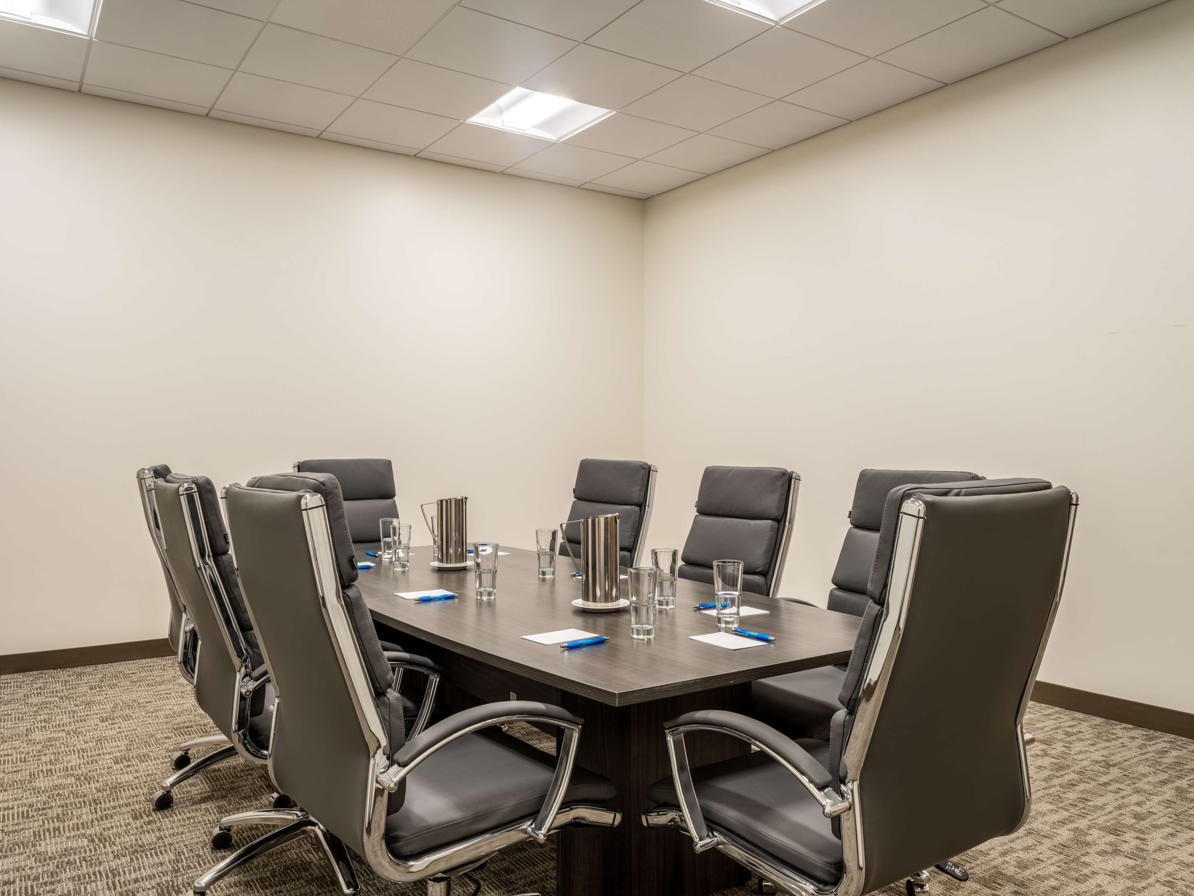 A conference room with a dark wooden table, six black chairs with chrome frames, and a carpeted floor.