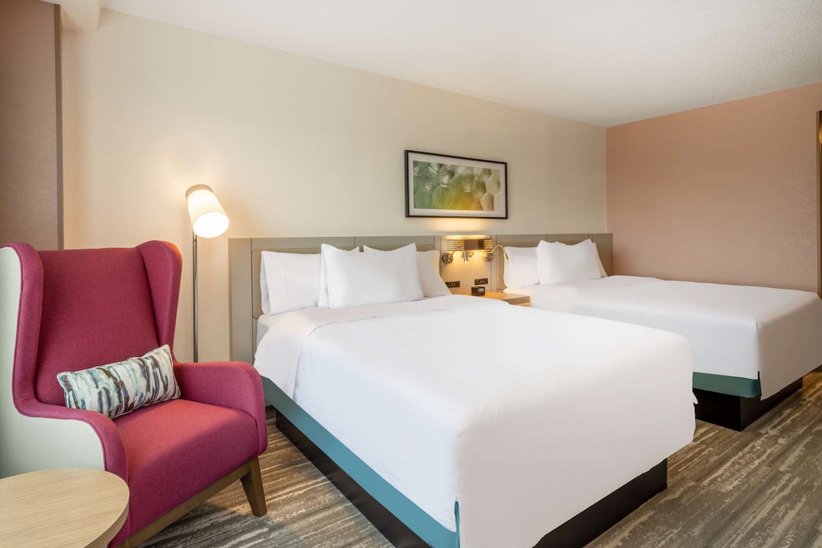 A hotel room with two beds, a red armchair, a small table, and a wall-mounted lamp.