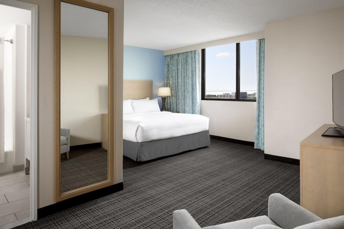 Ein Hotelzimmer mit Bett, Schreibtisch, Stuhl, einem großen Spiegel und einem Fenster mit Aussicht.
