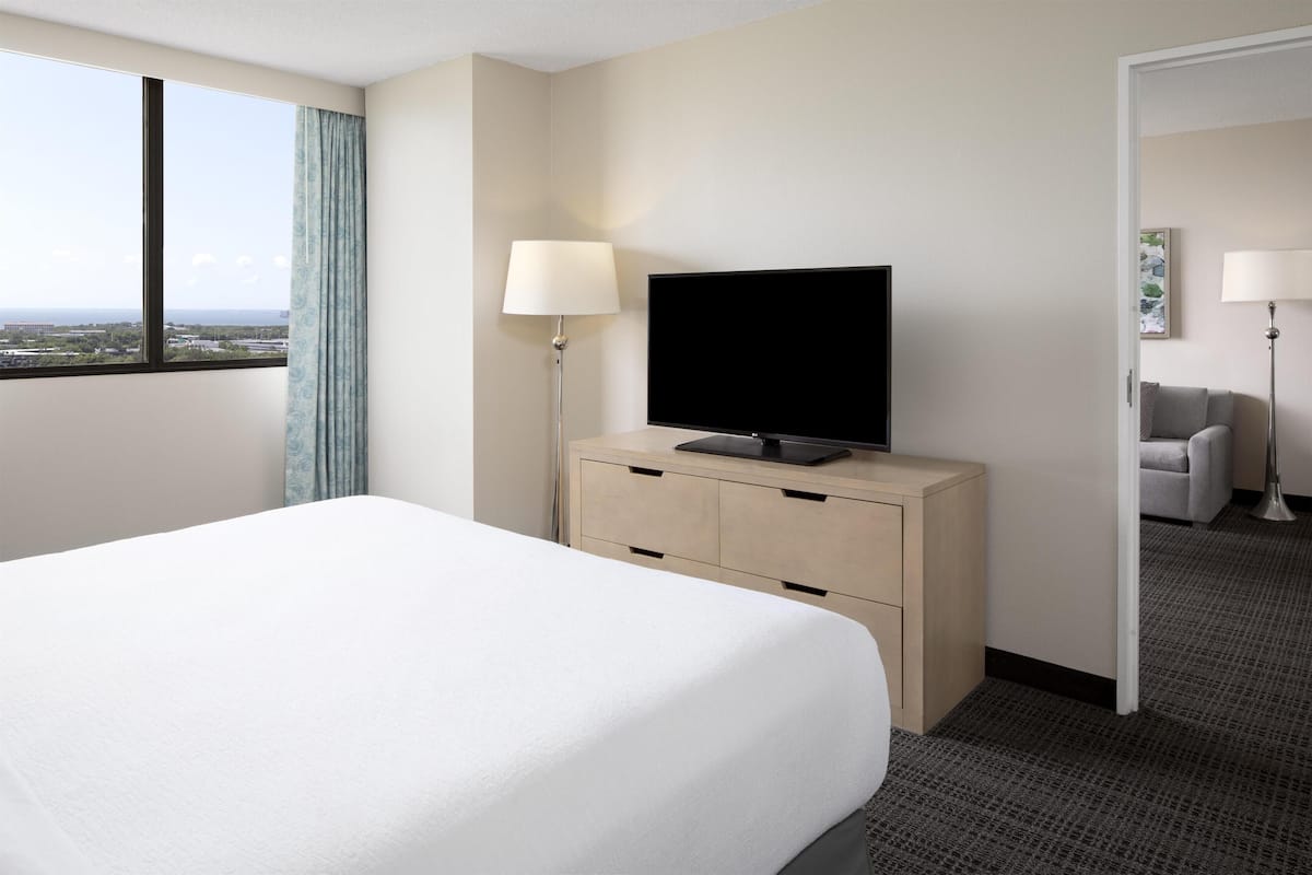 Ein Hotelzimmer mit Bett, einem Flachbildfernseher auf einer Holzkommode, einer Lampe und Blick auf die Stadt durch ein großes Fenster.