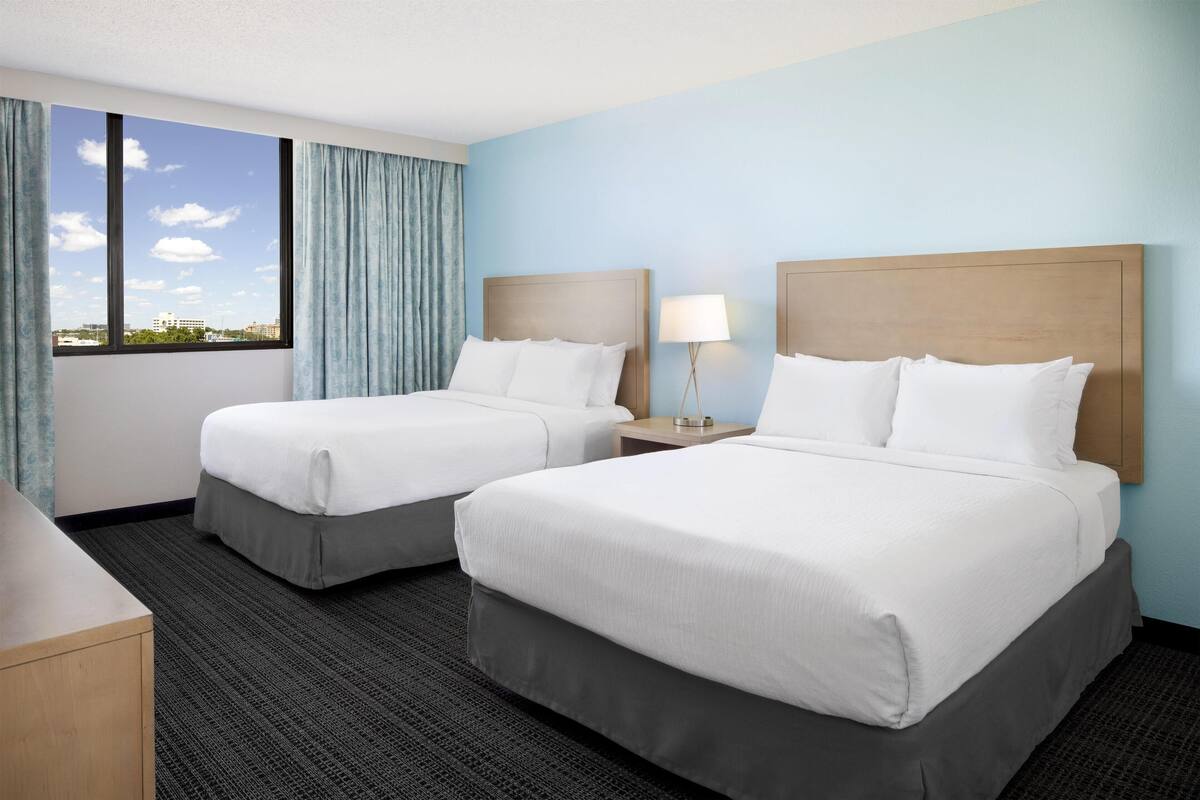 A hotel room with two beds, a nightstand with a lamp, a view of the city through large windows, and a carpeted floor.
