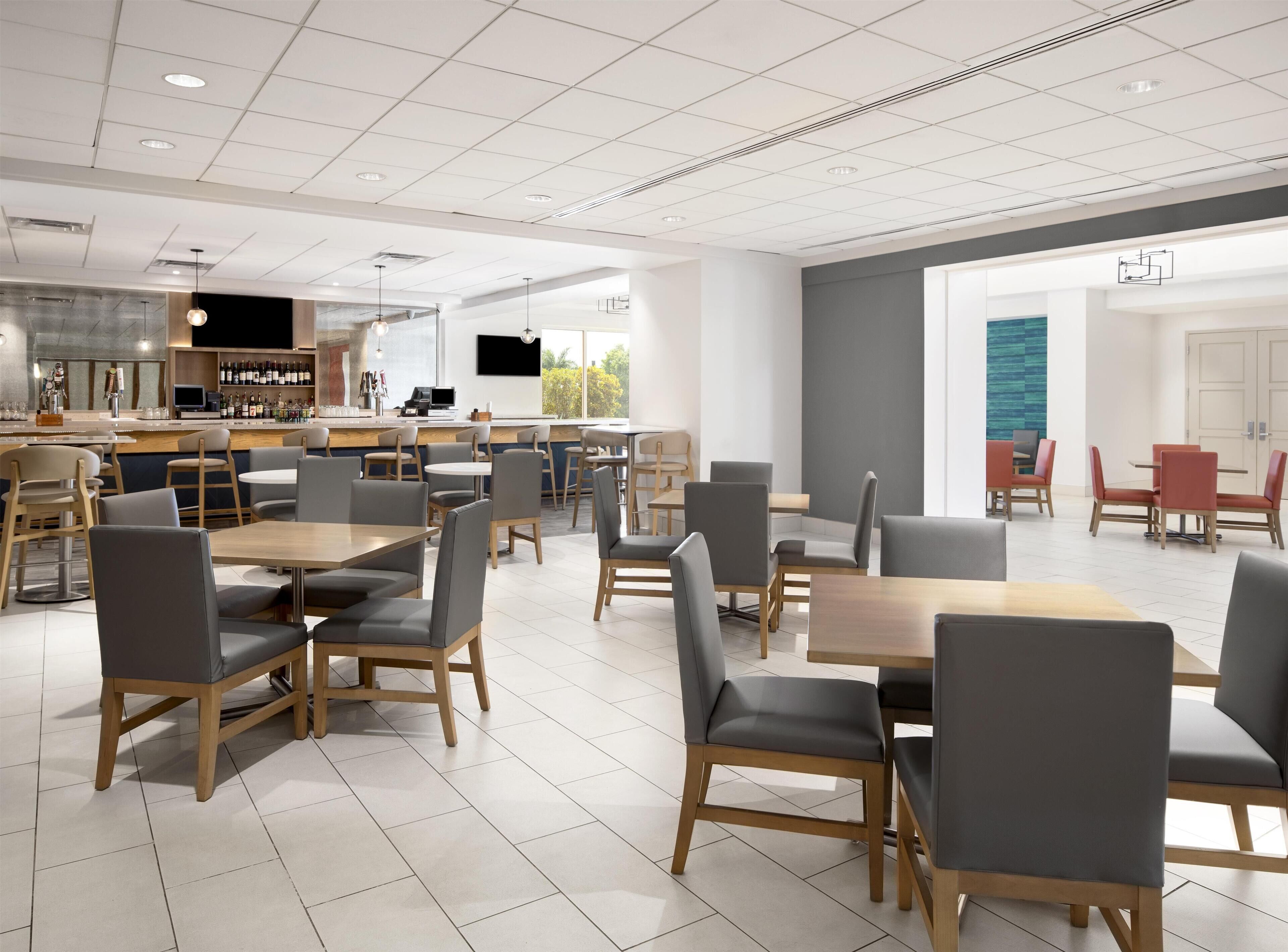 A modern cafe with multiple seating areas, a bar, and a large window providing natural light.