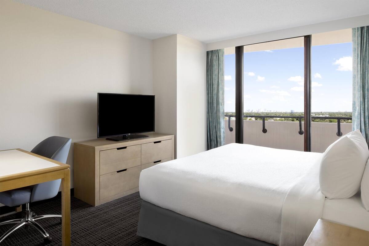 A hotel room with a bed, a desk, a television, and a large window with a city view.