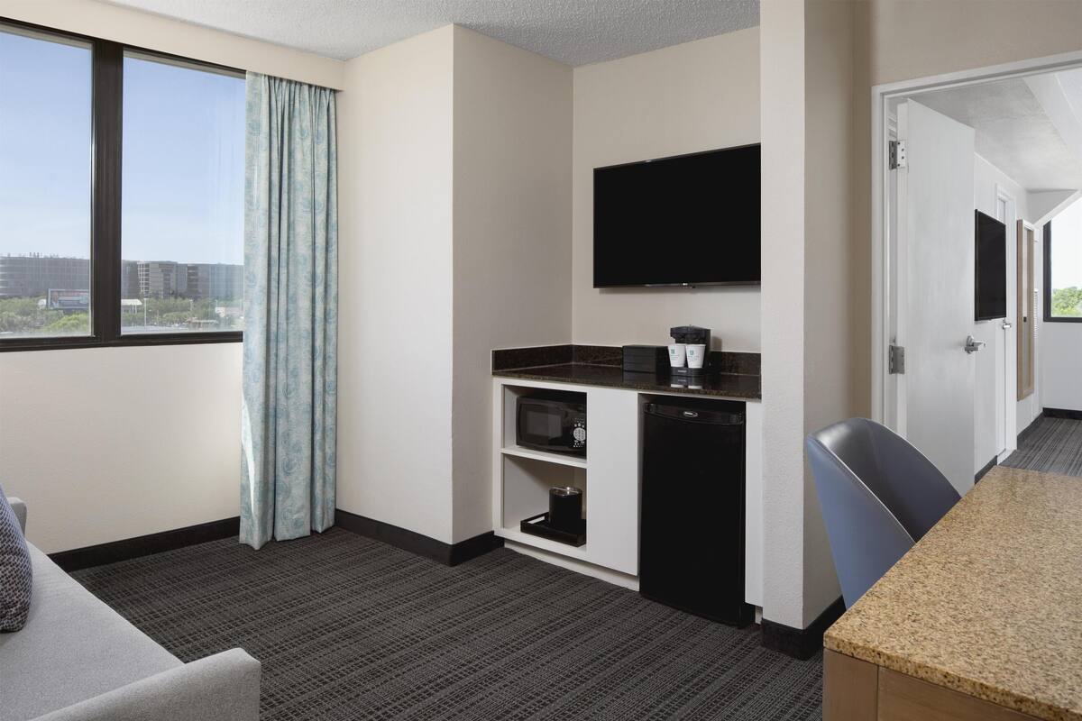 A hotel room with a large window, a flat-screen TV, a kitchenette, and a seating area.