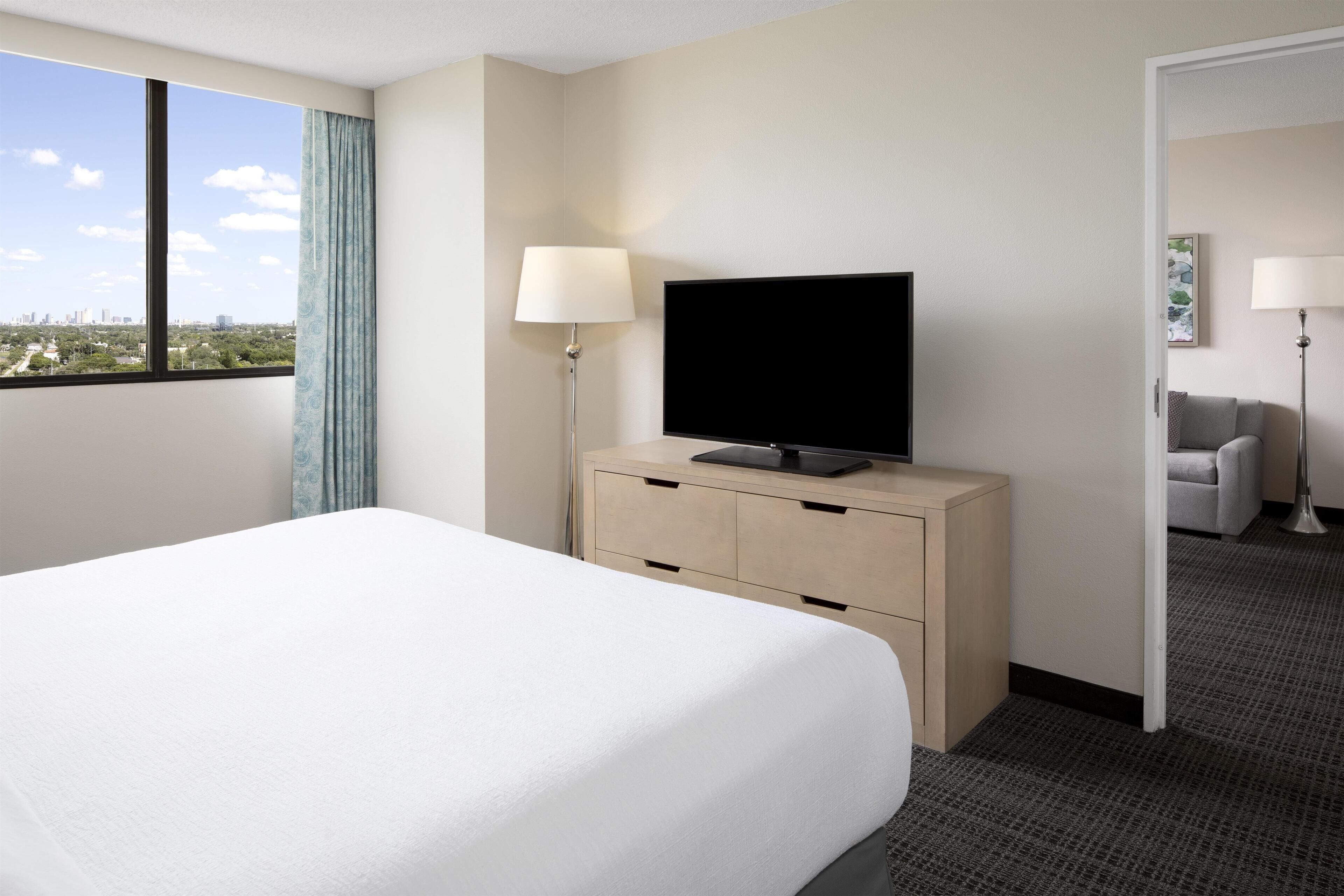Habitación de hotel con cama, un televisor de pantalla plana sobre un aparador de madera, una lámpara y vistas de la ciudad a través de un ventanal grande.