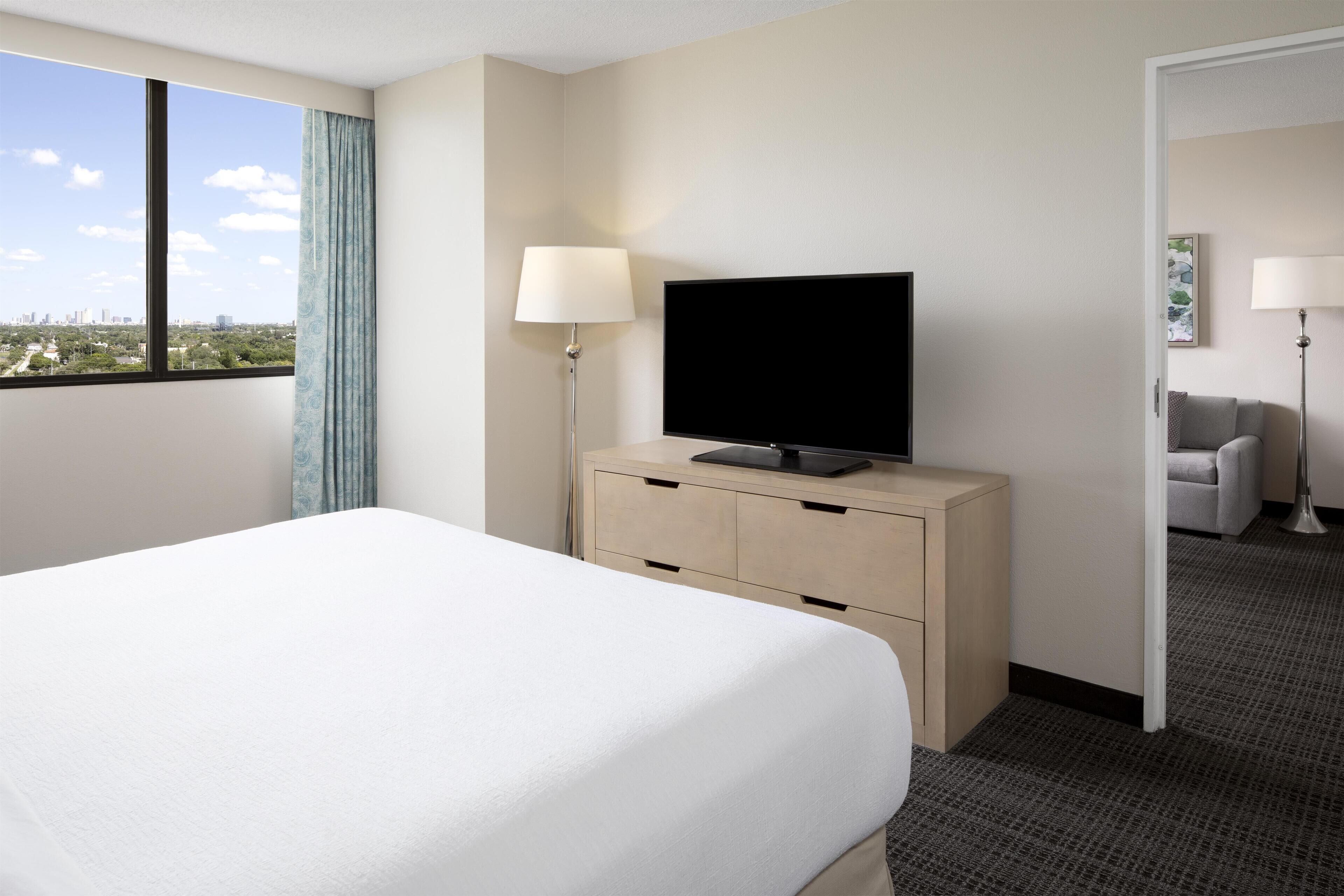 A hotel room with a bed, a TV on a wooden dresser, a lamp, and a view of the city.