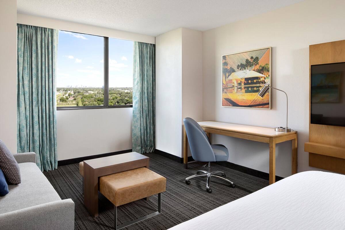 A hotel room with a bed, a desk, a chair, a bench, a large window with curtains, and a painting on the wall.