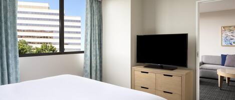 Una habitación de hotel con cama, televisor sobre un mueble de madera, ventana con cortinas y vista a un edificio.