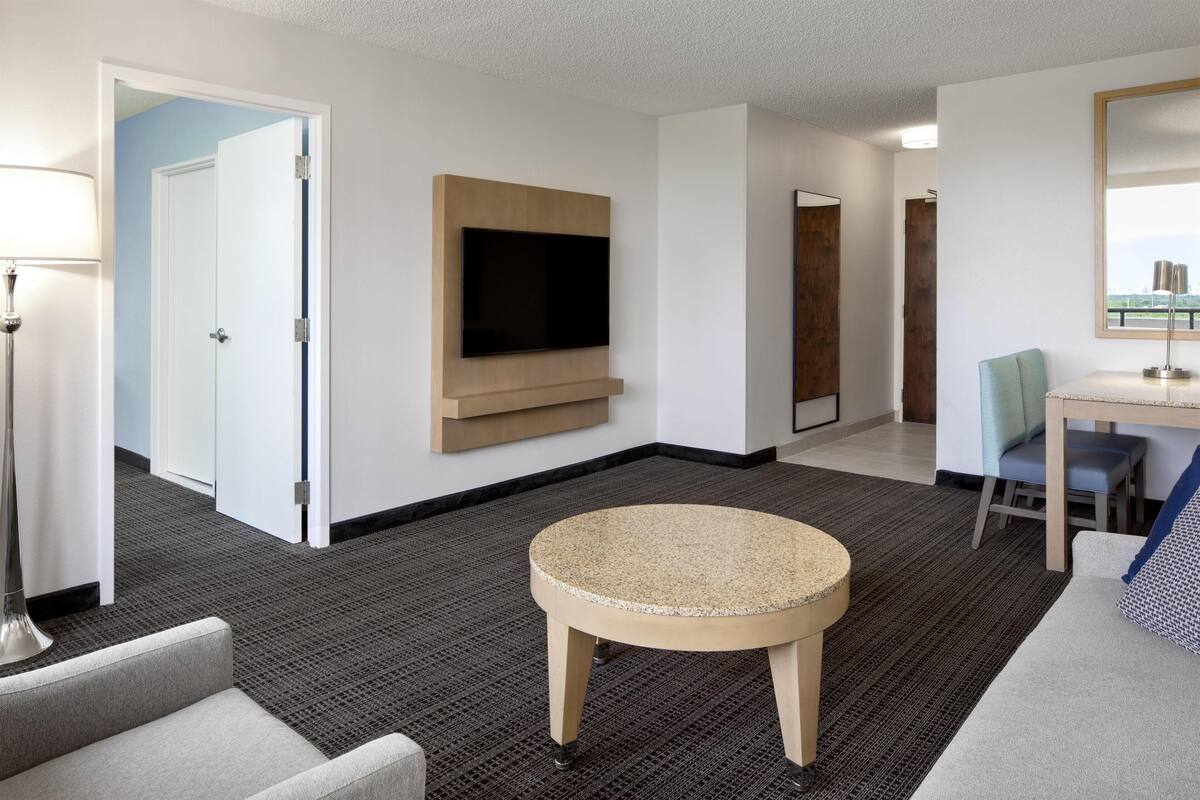A hotel room with a flat-screen TV mounted on the wall, a round table, a sofa, and a desk with a chair.