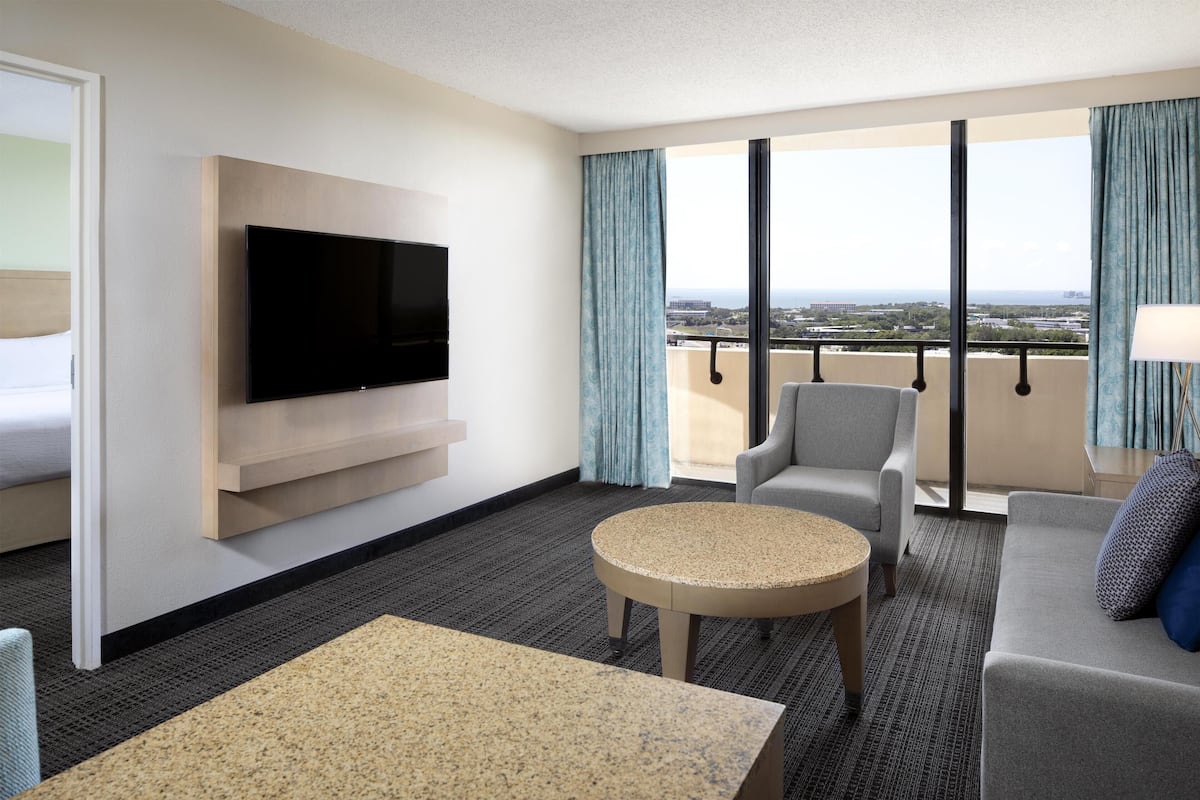 Ein modernes Hotelzimmer mit einem an der Wand montierten Flachbildfernseher, einem runden Couchtisch, einer Couch und einem Balkon mit Blick auf die Stadt.