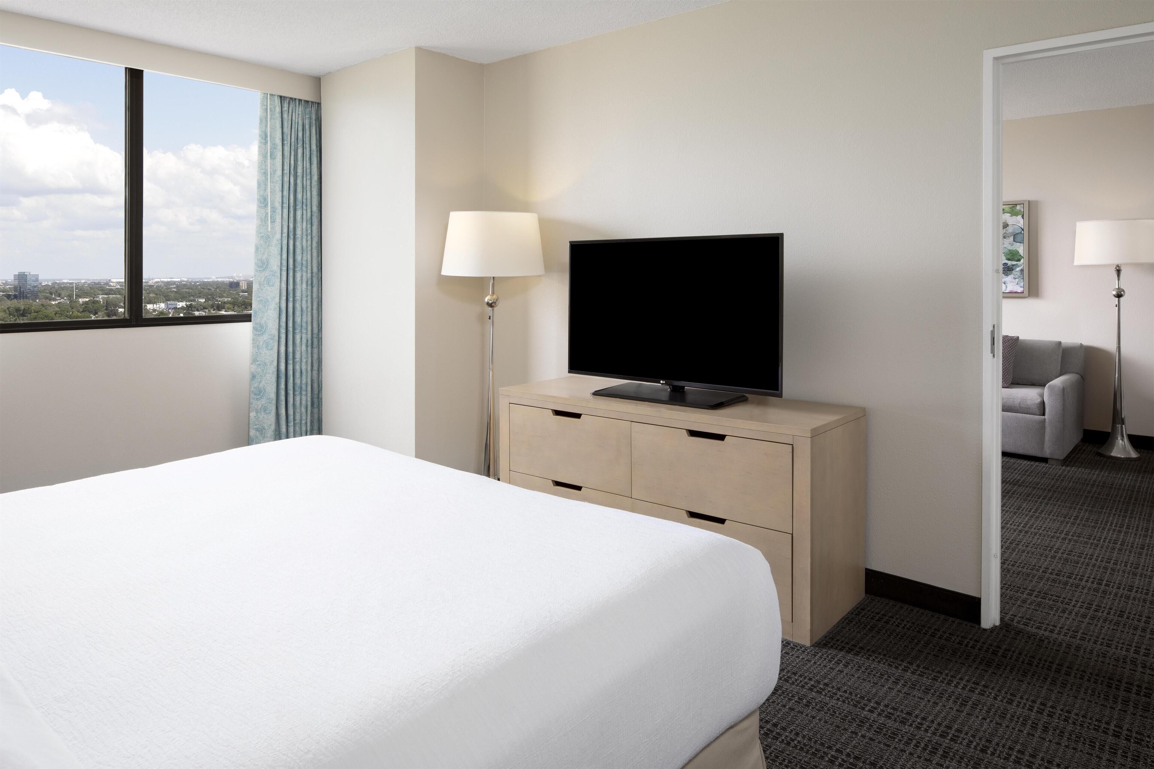 A hotel room with a bed, a TV on a wooden dresser, a lamp, and a view of the city through a window.