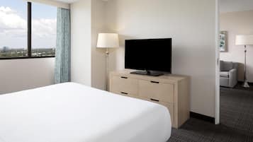 A hotel room with a bed, a TV on a wooden dresser, a lamp, and a view of the city through a window.