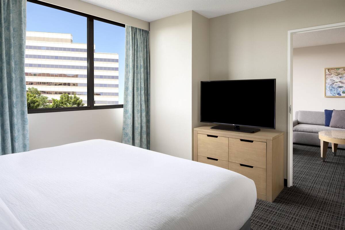 A hotel room with a large bed, a television on a wooden cabinet, and a view of a building through a window with curtains.