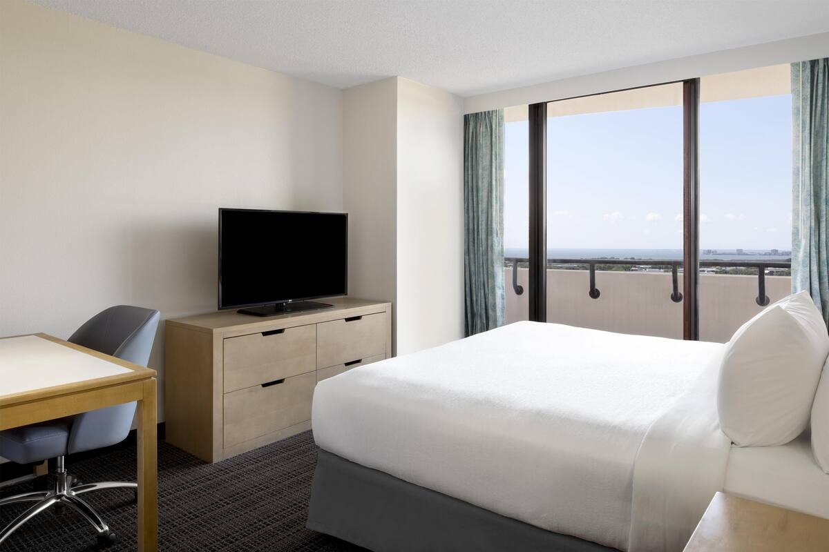 A hotel room with a bed, a desk, a television, and a view of the city through large windows.