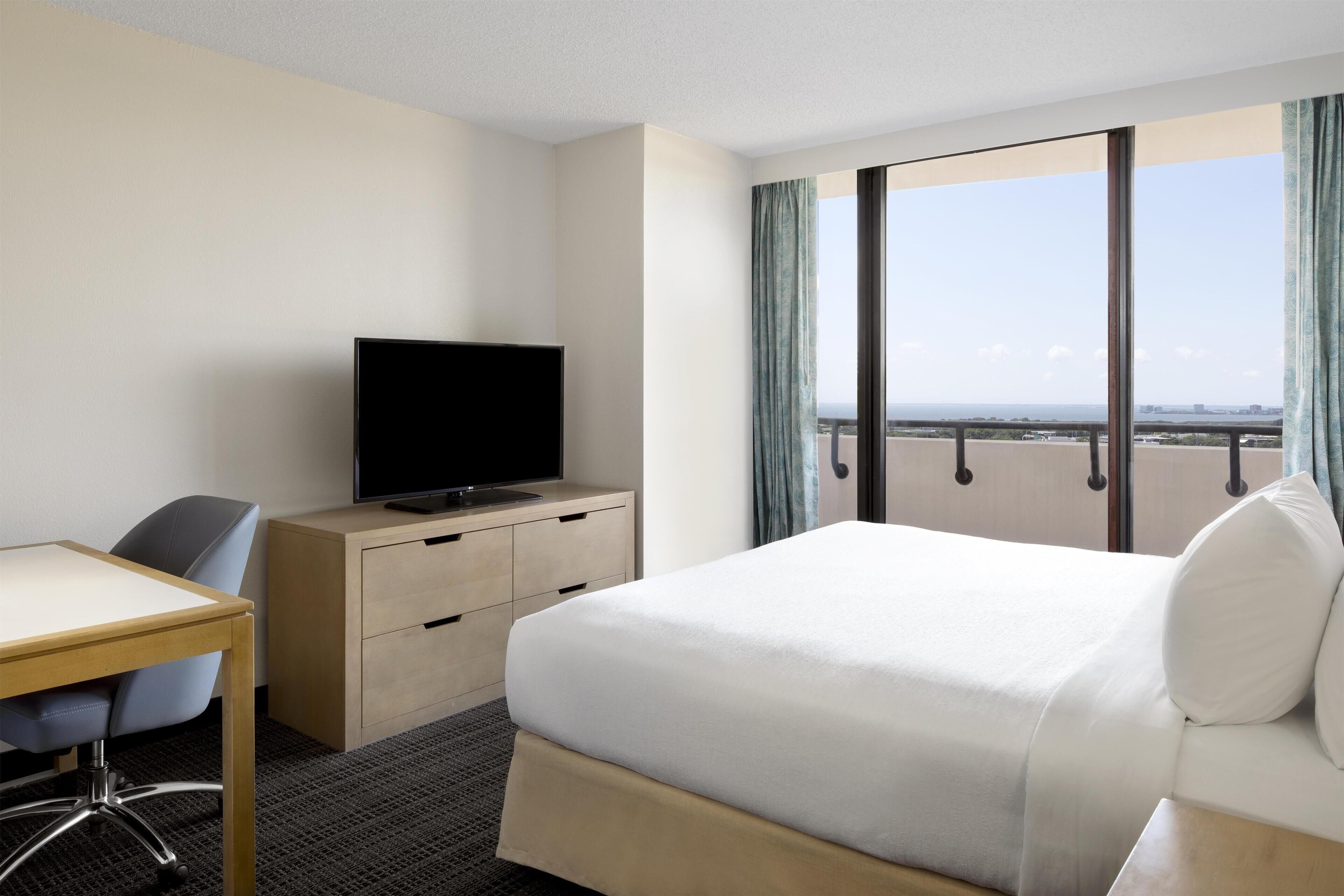 Habitación de hotel con cama, escritorio, televisor y vistas a la ciudad a través de amplios ventanales.