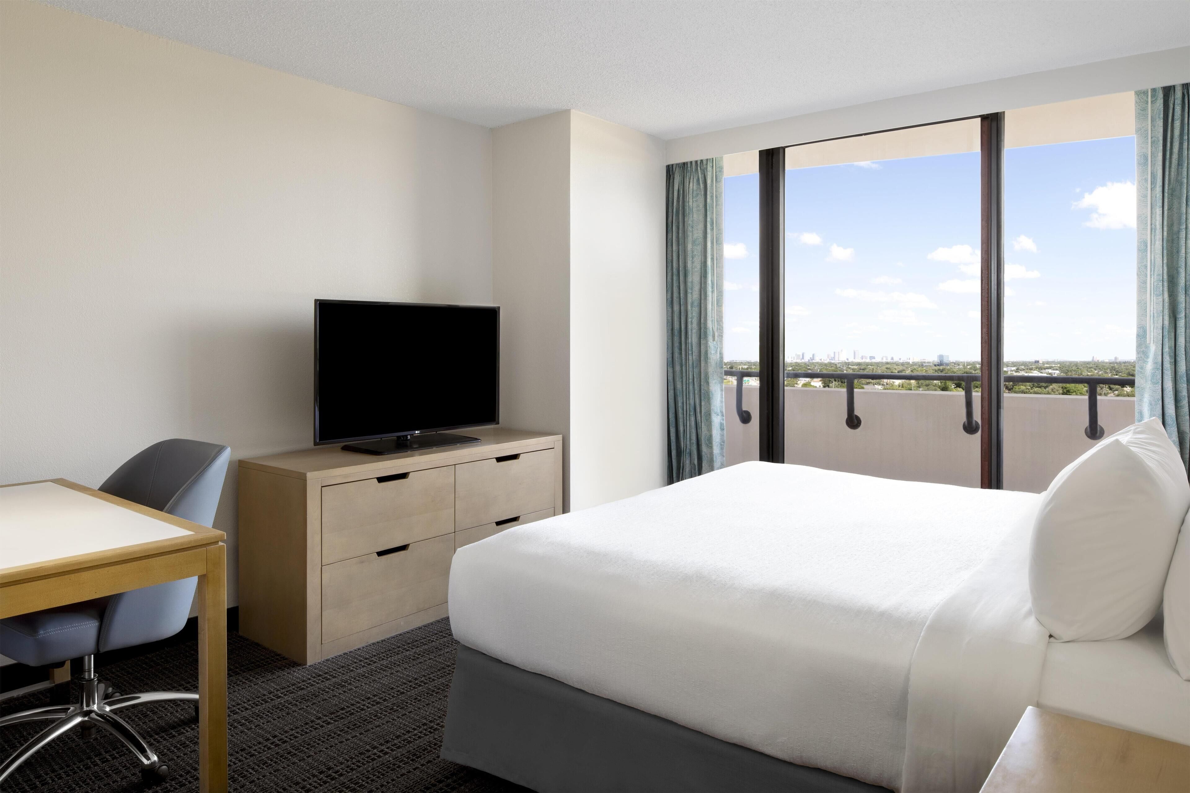 Une chambre d’hôtel équipée d’un lit, d’un bureau, d’une télévision et d’une grande fenêtre donnant sur la ville.