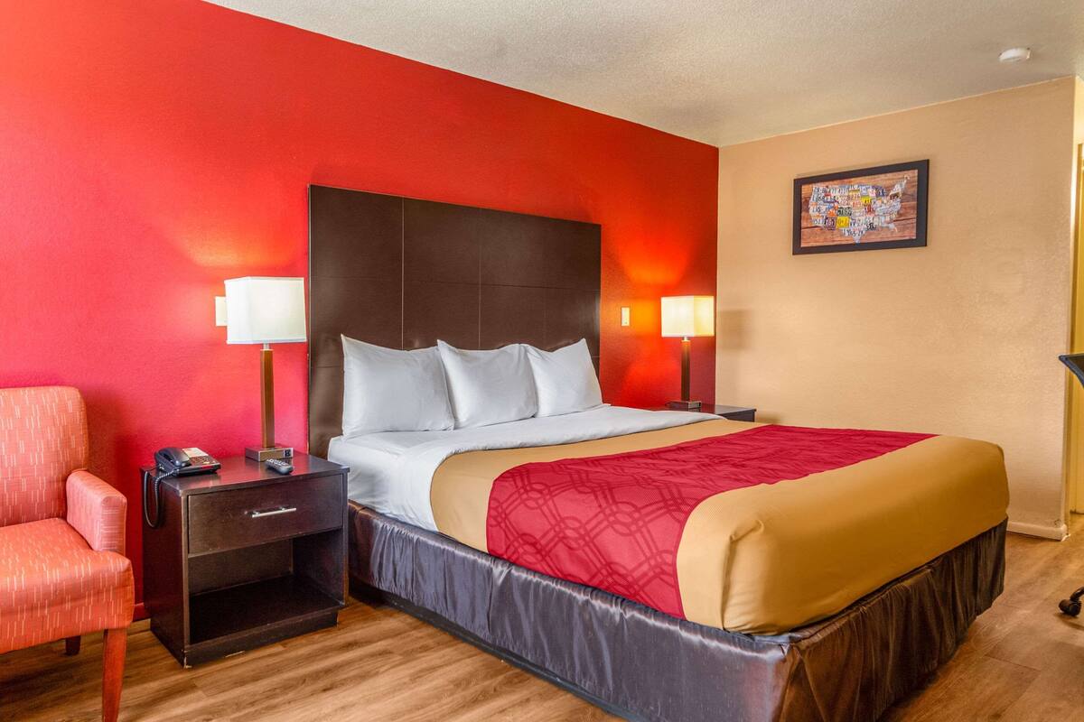 A hotel room with a red wall, a large bed with a red and beige bedspread, two bedside lamps, a nightstand with a phone, and a red armchair.