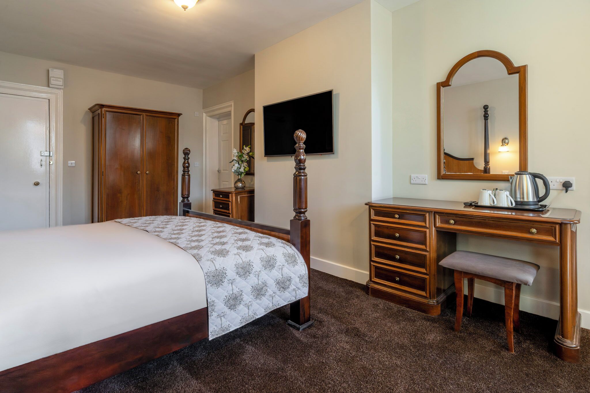 A bedroom with a bed, a wooden dresser, a desk with a mirror, and a television.