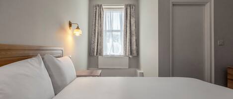 A hotel room with a bed, a nightstand, a wall-mounted lamp, a window with curtains, and a door.