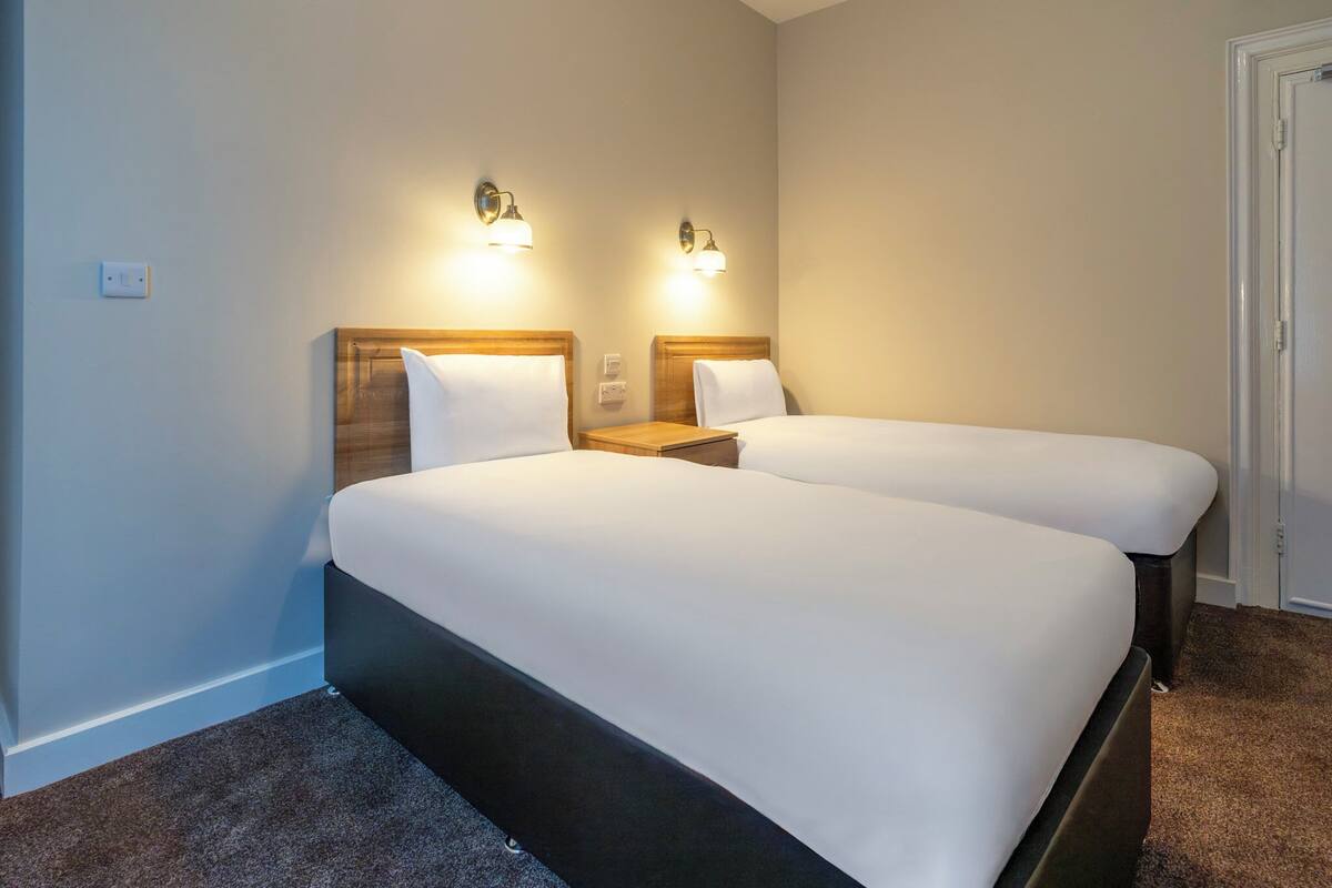 A hotel room with two beds, wooden headboards, bedside tables, and wall-mounted lamps.