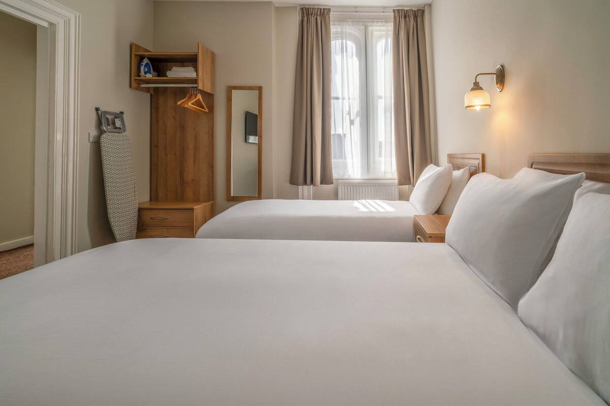 A hotel room with two beds, a wooden wardrobe, a mirror, a radiator, and a wall-mounted light fixture.