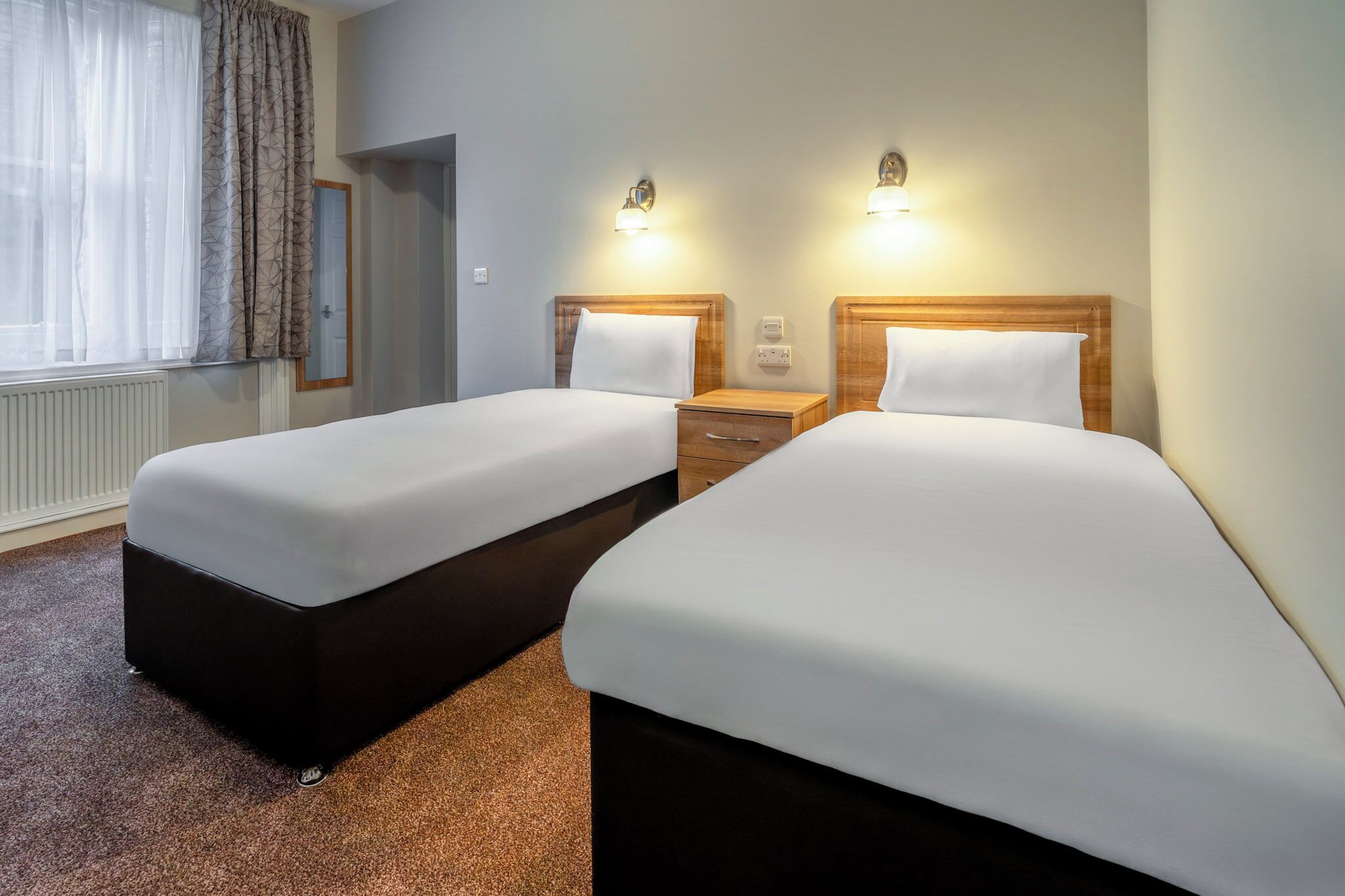A hotel room with two beds, a nightstand, a mirror, and two wall-mounted lights.