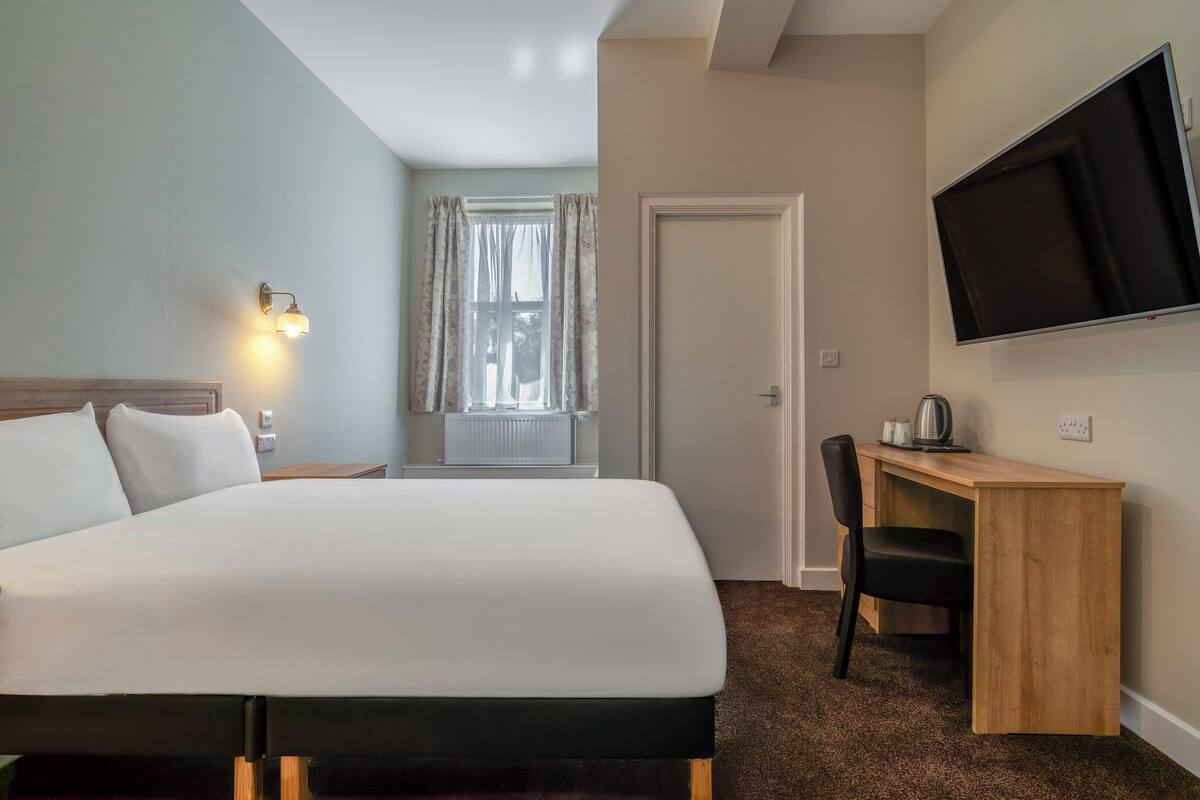 A hotel room with a bed, a desk with a chair, a television, and a window with curtains.
