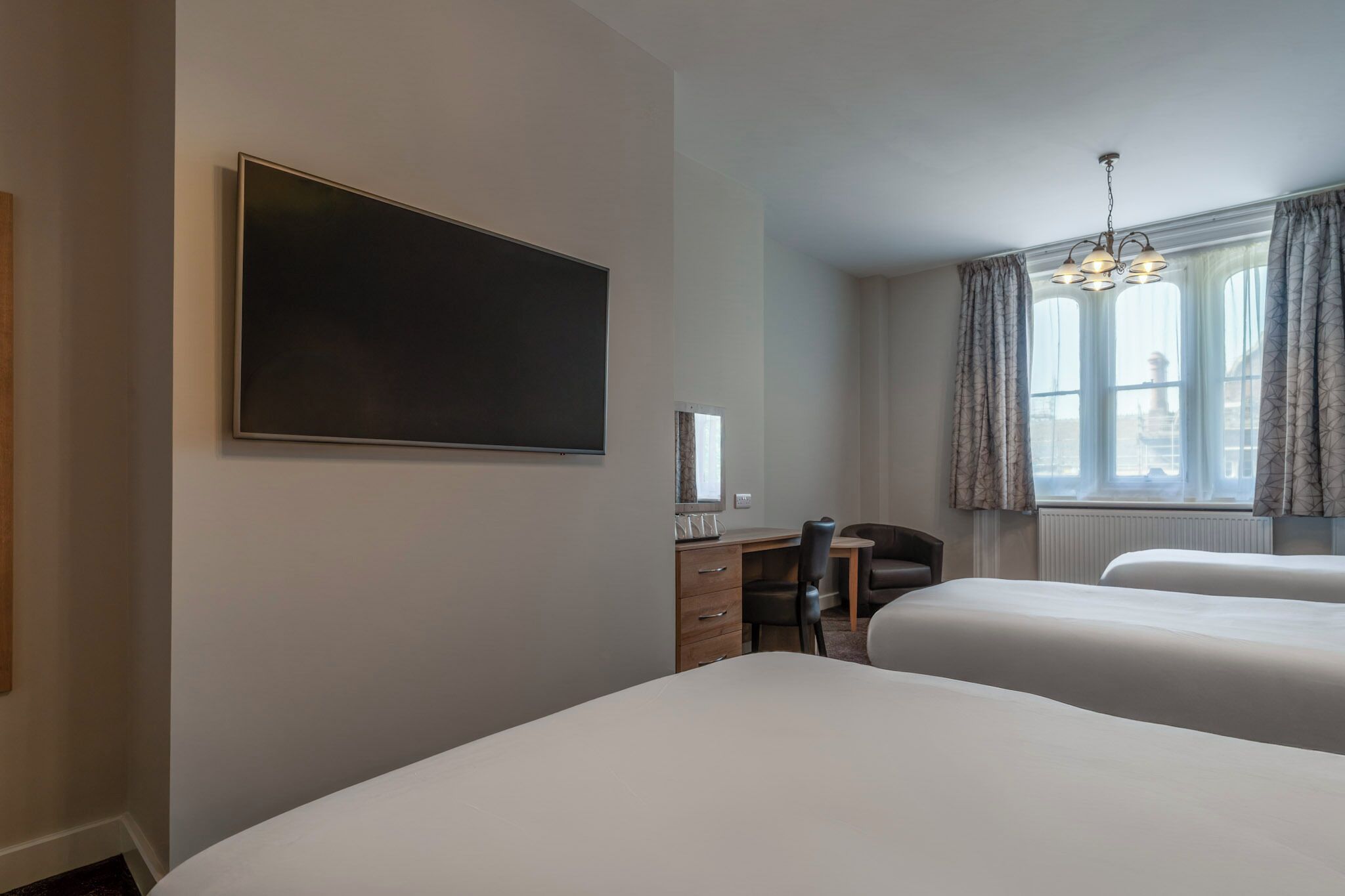 A hotel room with two beds, a desk with a chair, a flat-screen TV, and a window with curtains.