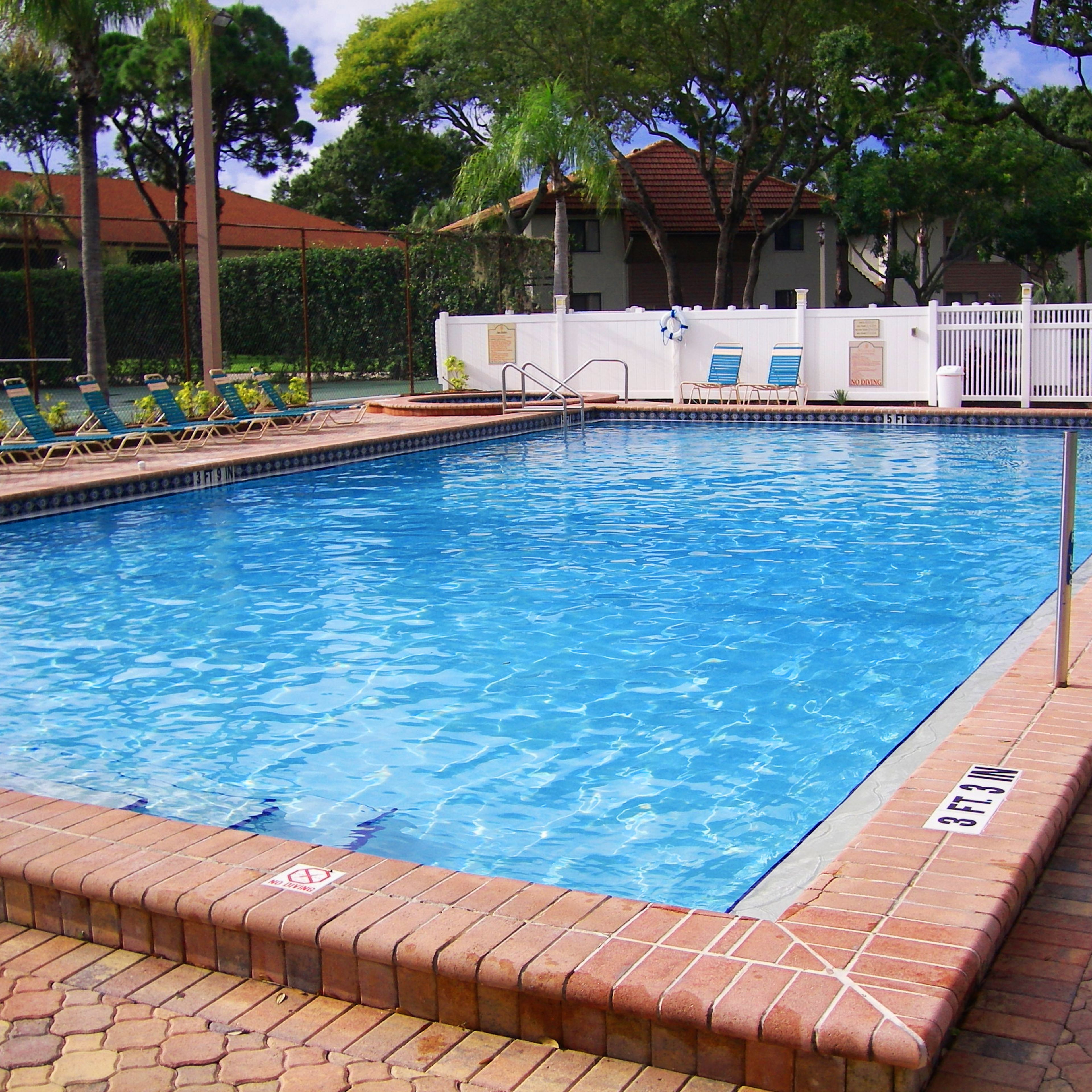A rectangular swimming pool with a brick edge, surrounded by a white fence, and equipped with lounge chairs and a lifeguard stand.