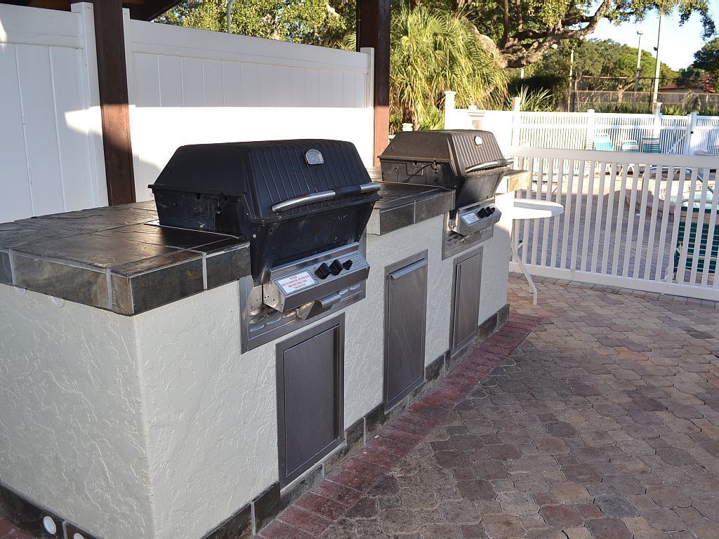 Ett utomhusområde med grill, en inbyggd ugn och en rökugn, omgivet av ett vitt staket och en pool.