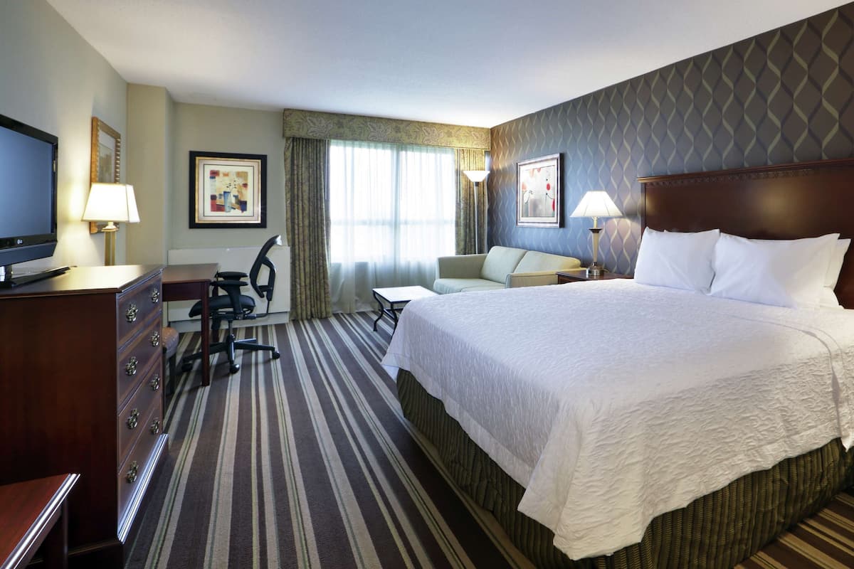 A hotel room with a large bed, a desk with a chair, a television, and a nightstand with a lamp.
