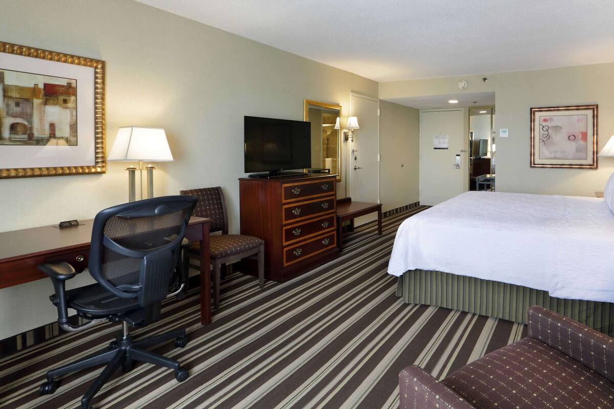 A hotel room with a bed, desk, chair, dresser, television, and lamps.