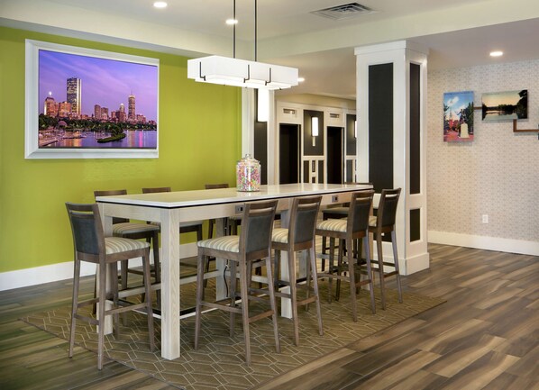 A modern dining area with a long table and chairs, a cityscape painting on the wall, and a patterned rug.