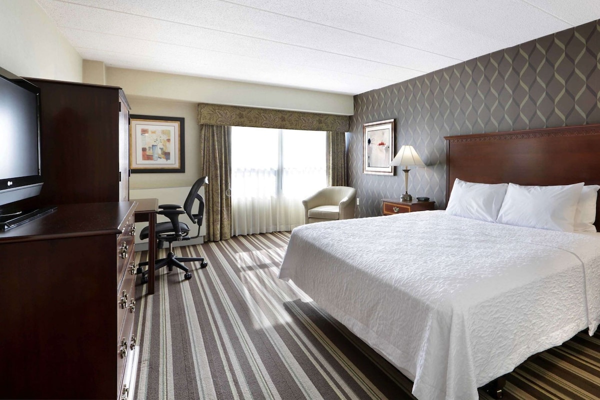 A hotel room with a large bed, a desk with a chair, a television, and a window with curtains.