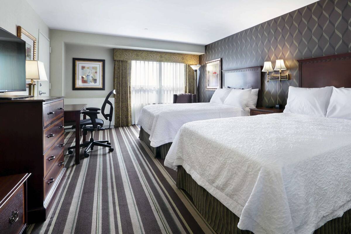 A hotel room with two beds, a desk, a chair, a TV, and a window with curtains.