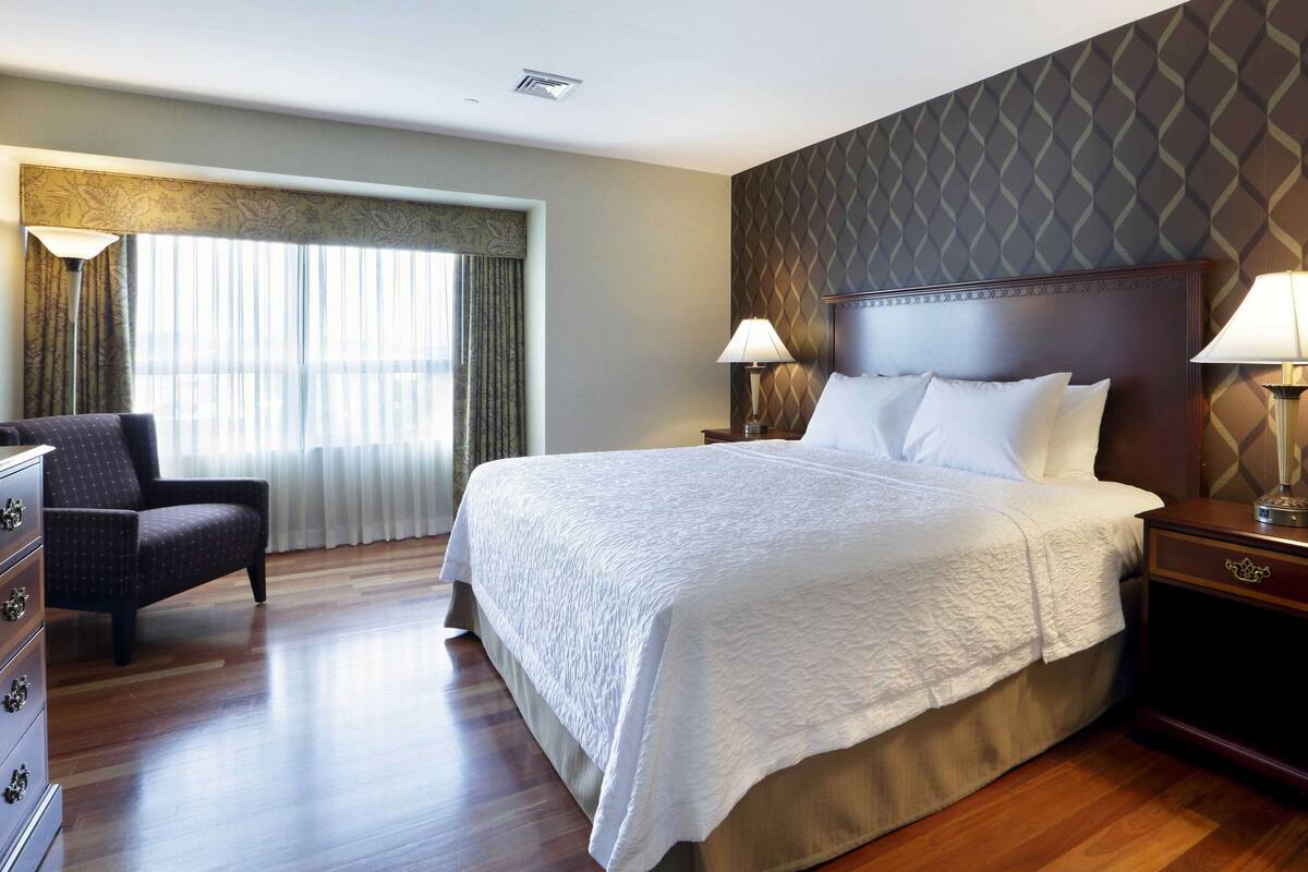 A hotel room with a large bed, two bedside lamps, a wooden nightstand, a chair, and a dresser.