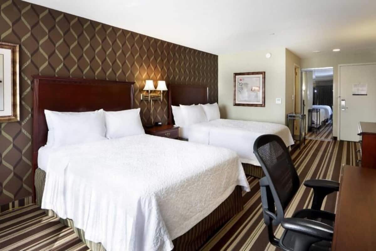 A hotel room with two beds, a desk, a chair, a TV, and a door leading to another room.